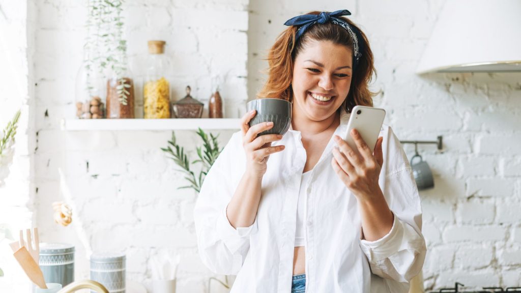 Una mujer sonriente en su cocina tomando una taza de café y consultando su teléfono.